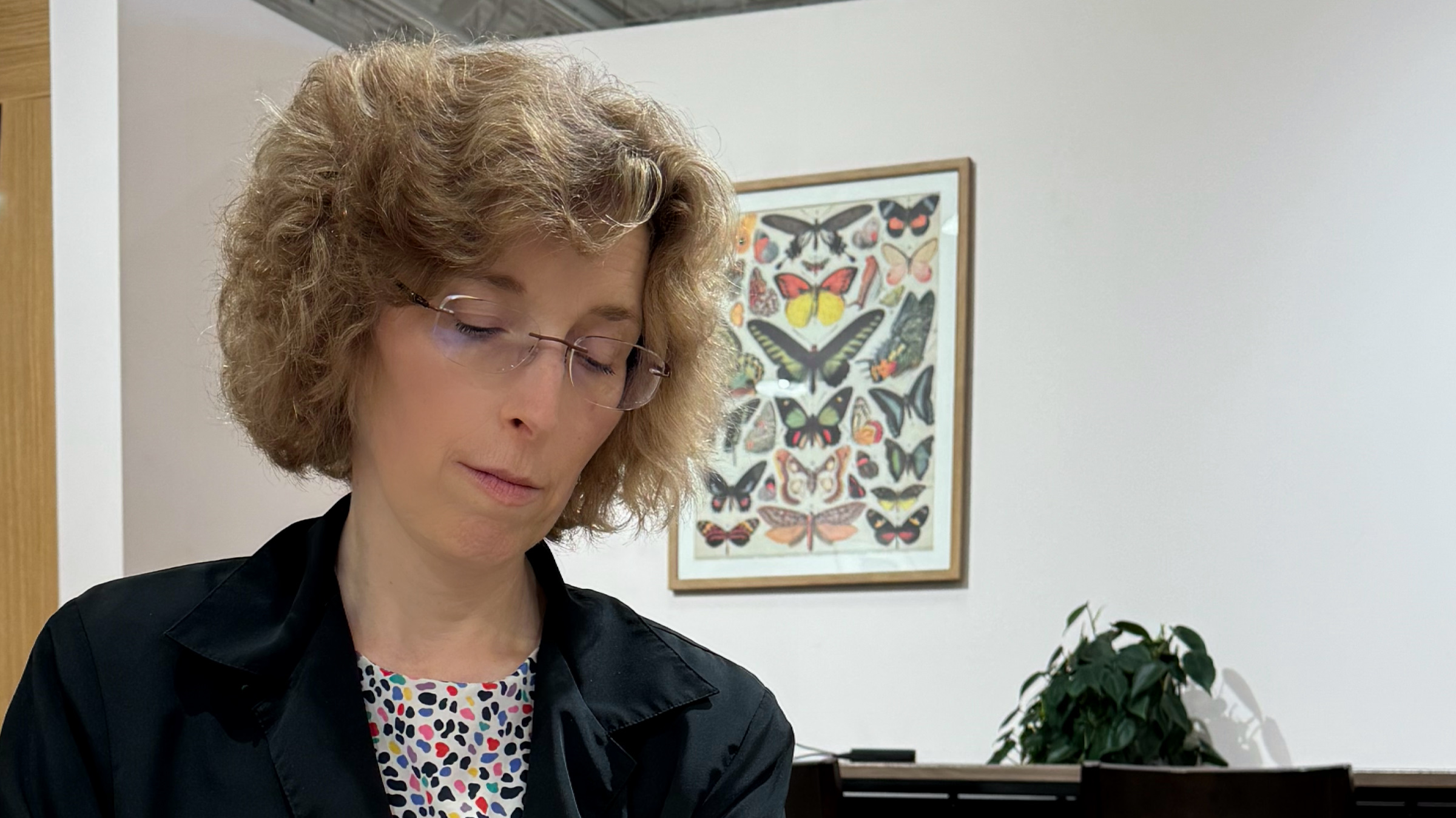 Kim in her glasses and a blue blazer taking notes with an framed poster of butterflies in the background.