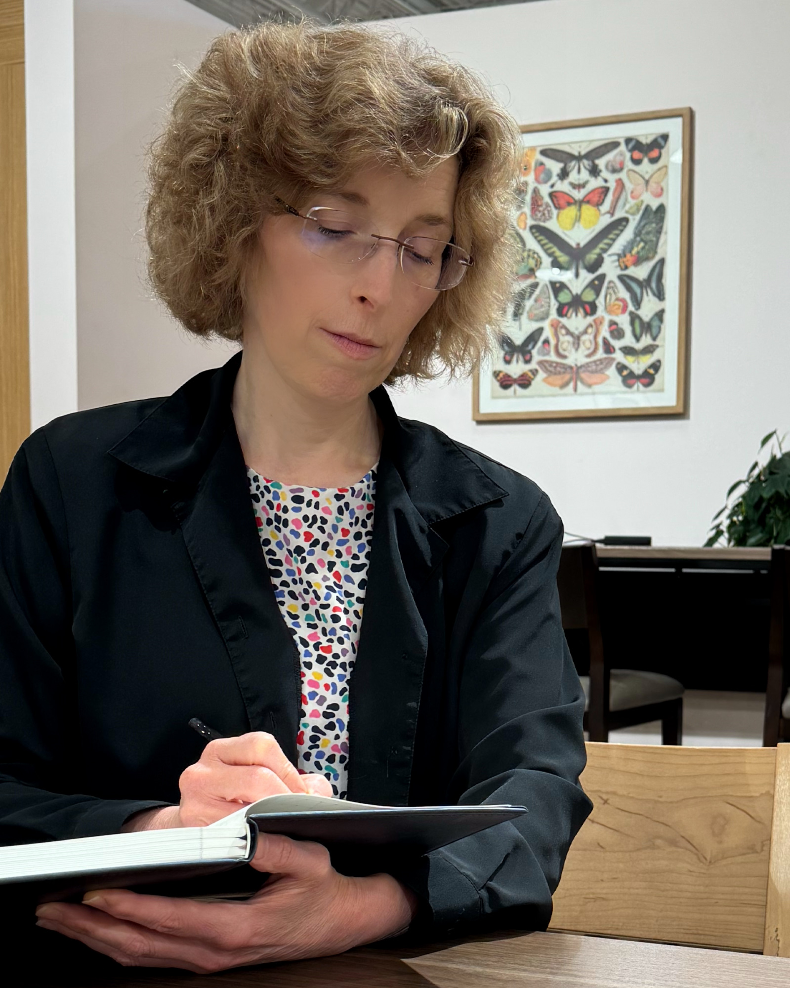 Kim in her glasses and a blue blazer taking notes with an framed poster of butterflies in the background.