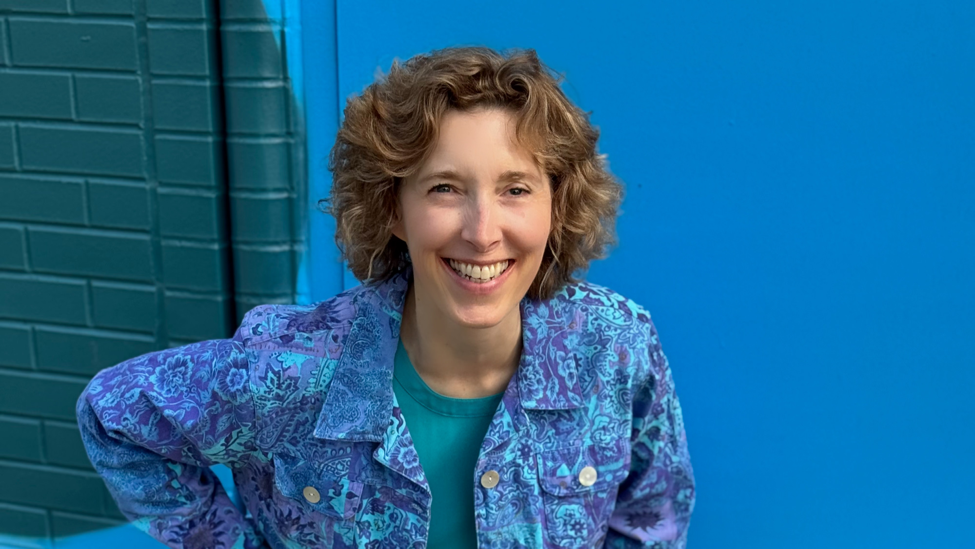 Kim smiling, in a blue and purple shirt, bent down in front of a brick wall with splashes of blue and green colors.