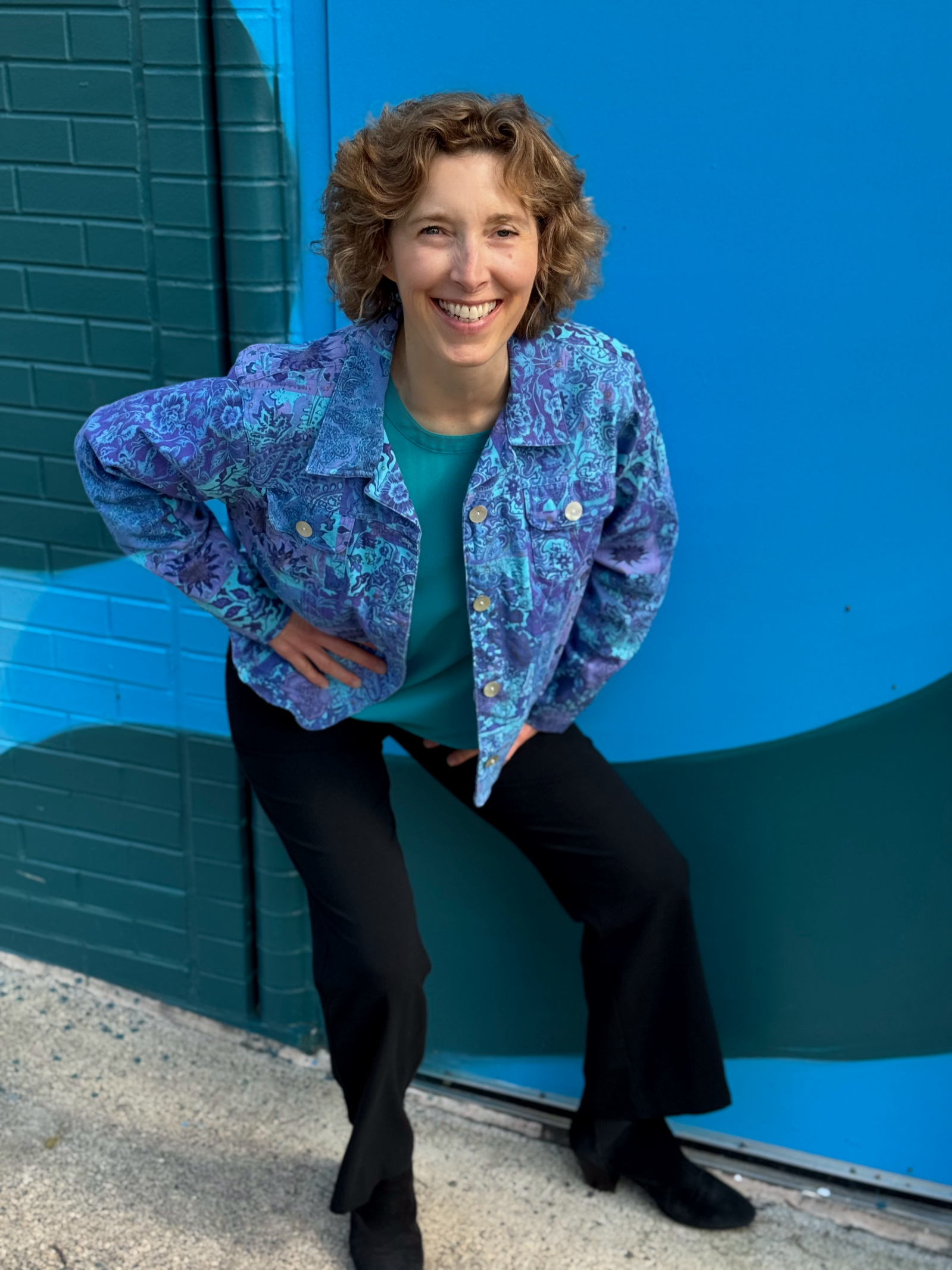 Kim smiling, in a blue and purple shirt, bent down in front of a brick wall with splashes of blue and green colors.