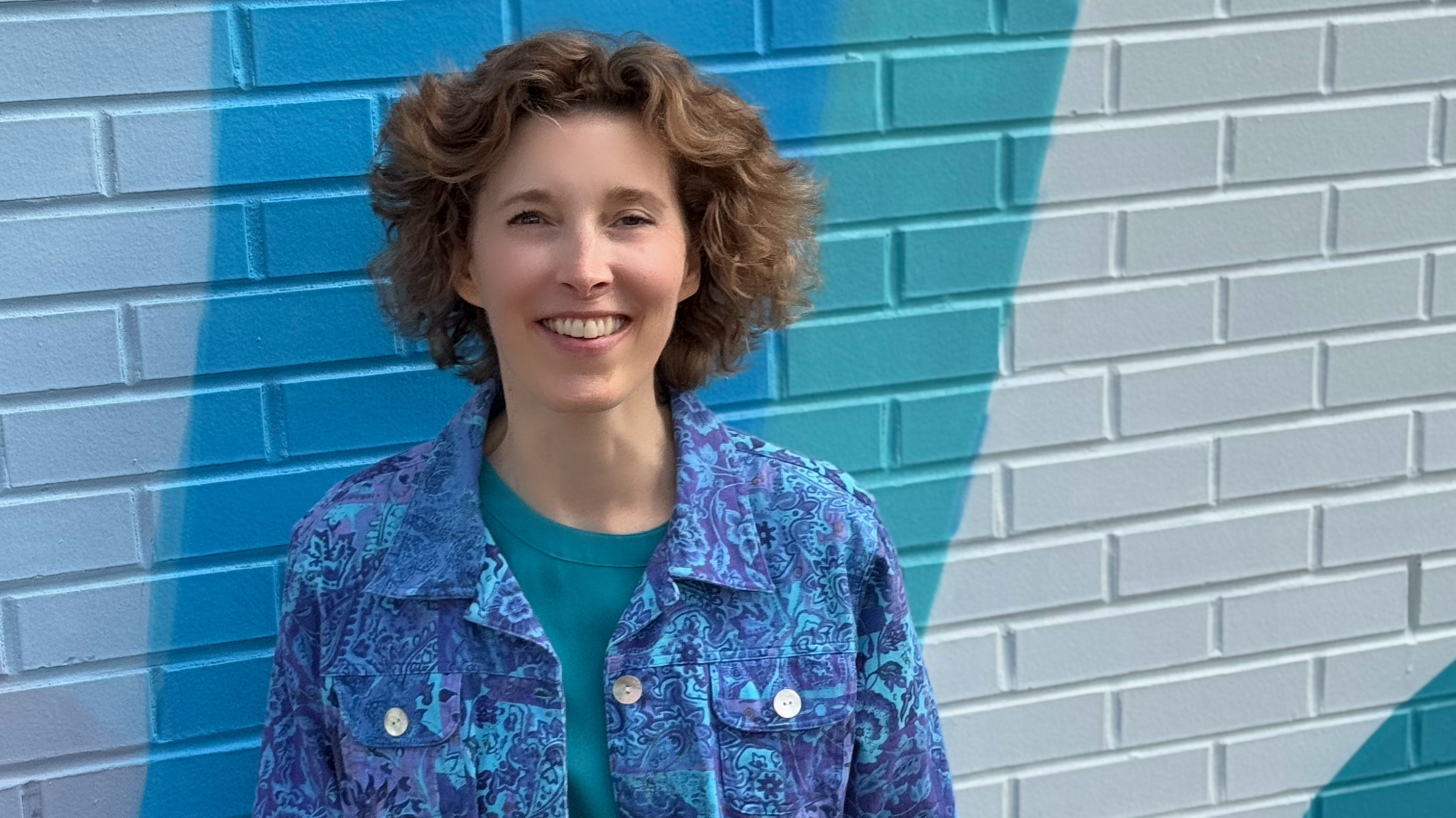 Kim smiling, in a blue and purple shirt, in front of a brick wall with diagonal of gray, blue, turquoise, and white colors.