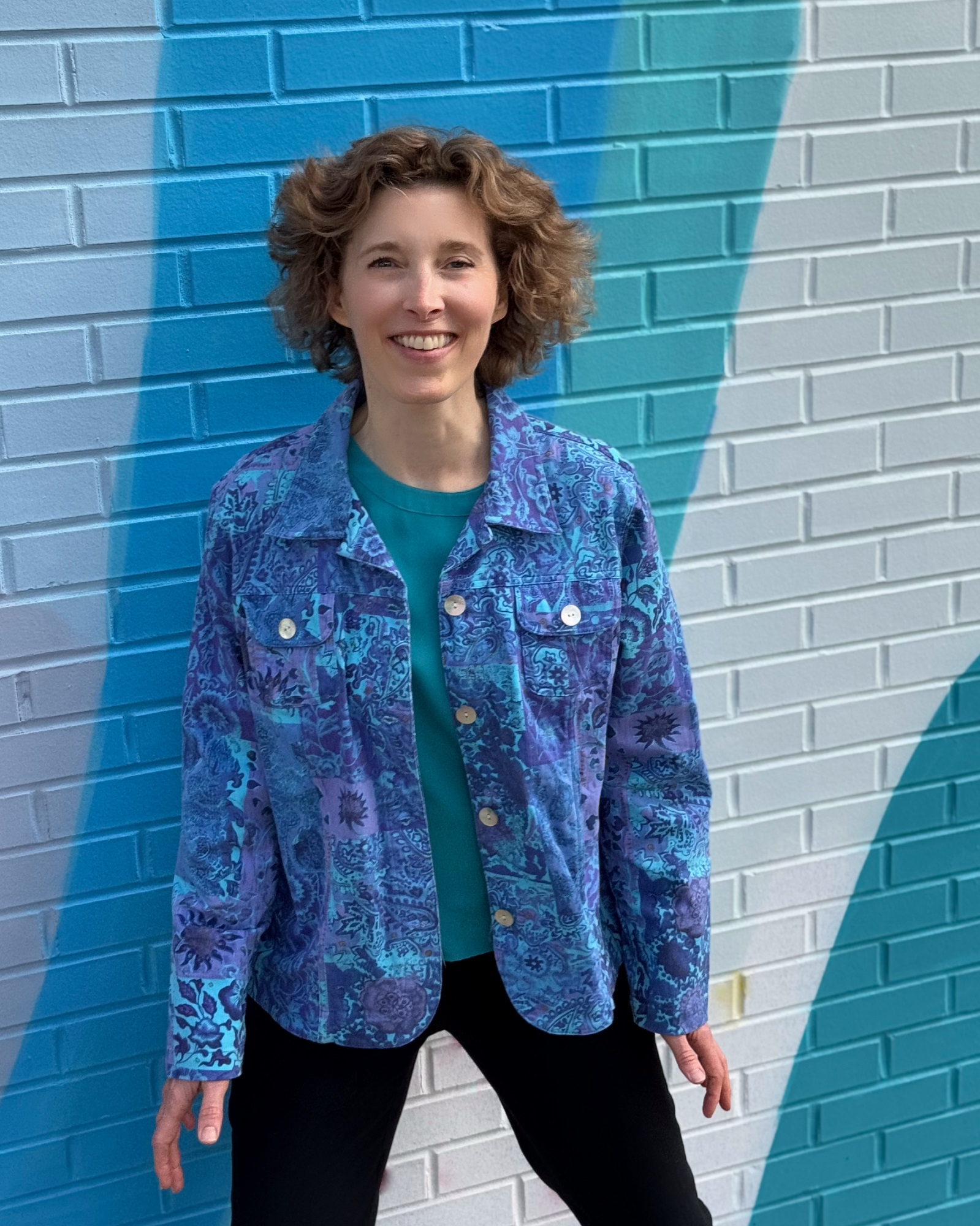 Kim smiling, in a blue and purple shirt, in front of a brick wall with diagonal of gray, blue, turquoise, and white colors.