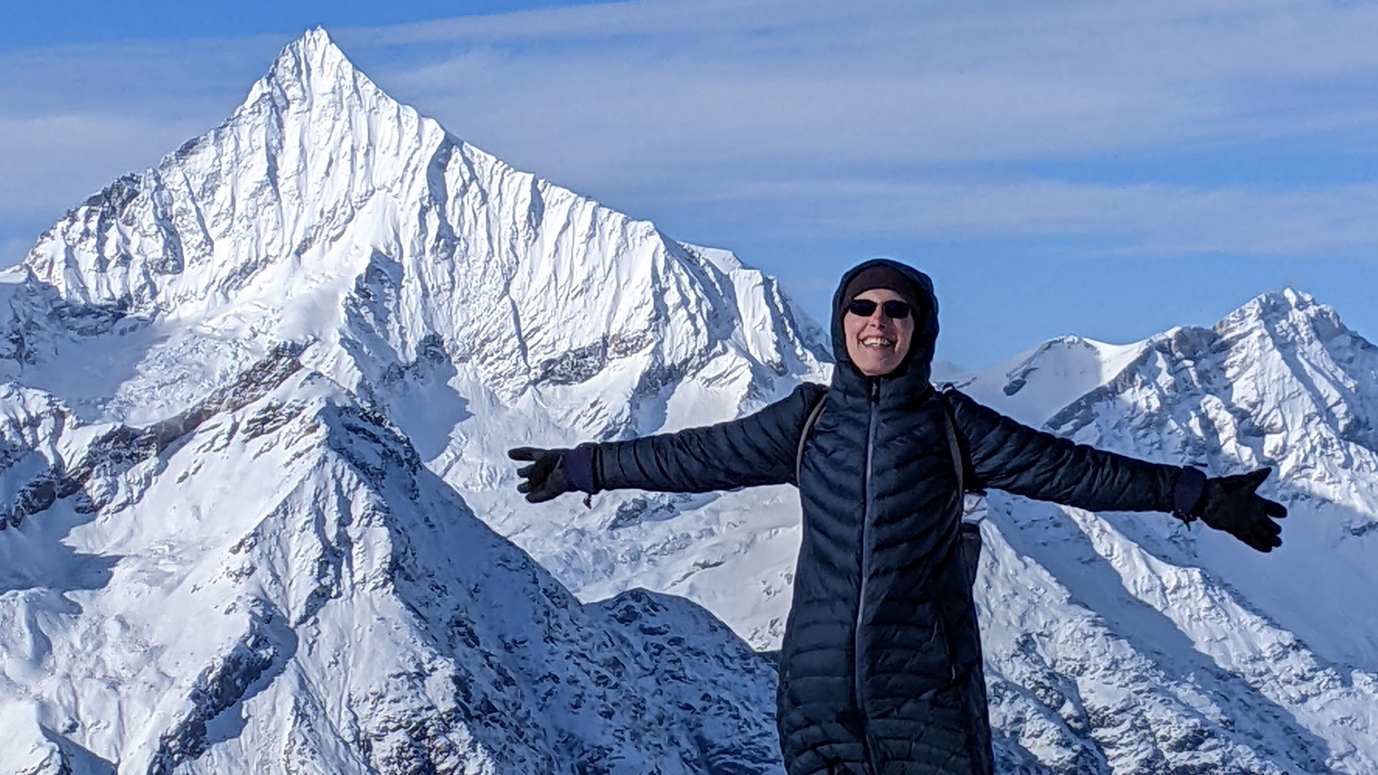 Kim in sunglasses and her winter parka, arms spread wide and smailing from a snow covered Gornergrat Switzerland with the Alps in the background.