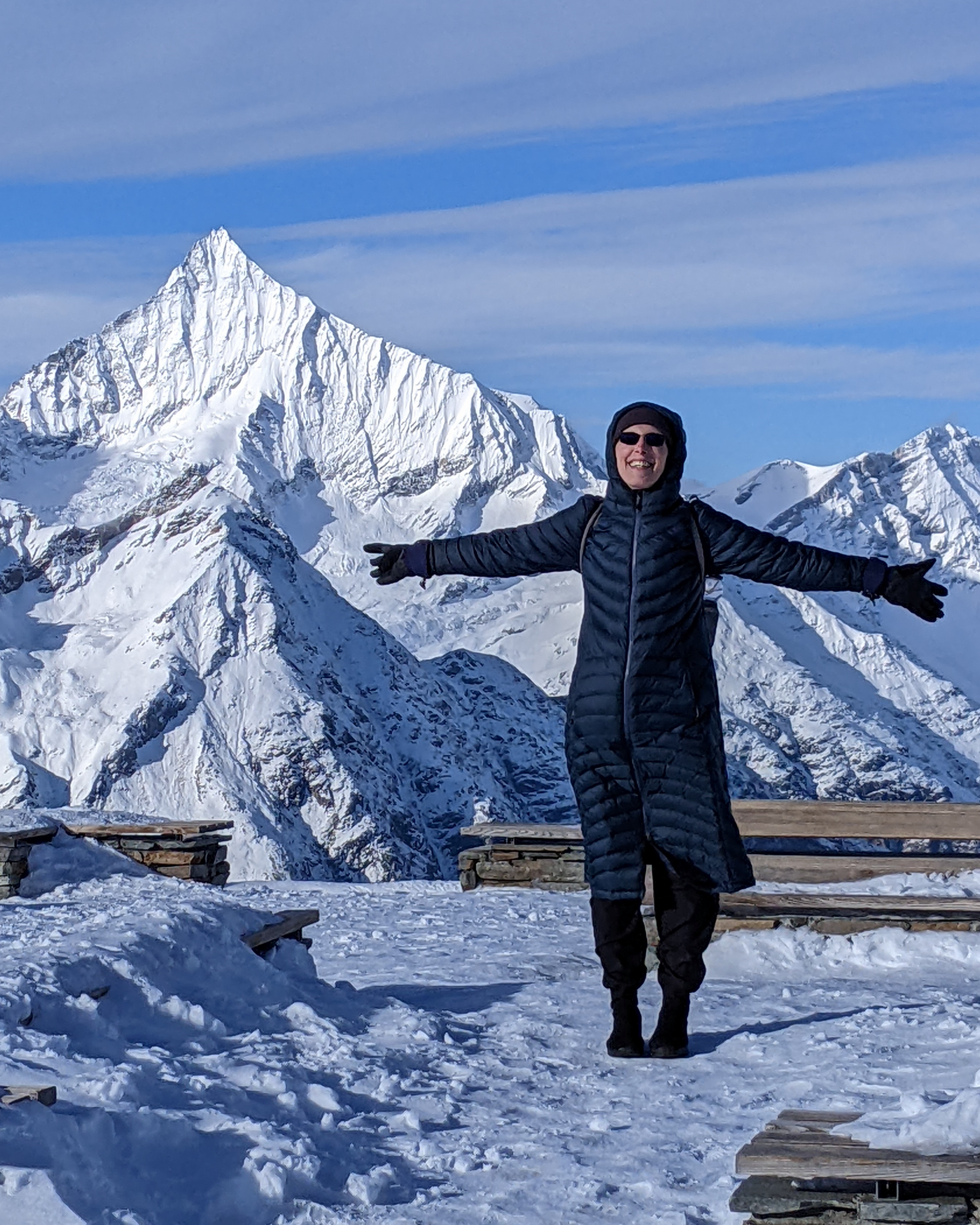 Kim in sunglasses and her winter parka, arms spread wide and smailing from a snow covered Gornergrat Switzerland with the Alps in the background.